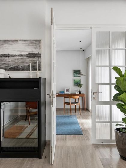 A peek into a study from the hallway. The clean white walls, minimalist furniture, and a large fiddle-leaf fig plant create a space that is bright, calm, and conducive to focus.