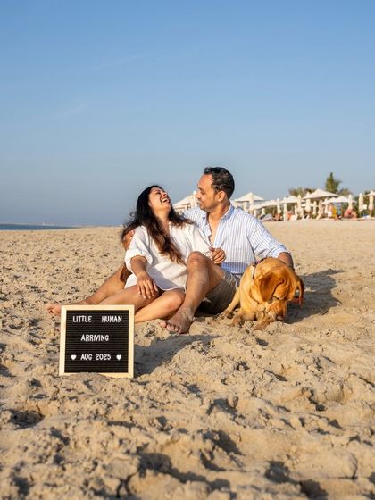 A joyful pregnancy announcement on the sunny beaches of Dubai. Astro the Labrador relaxes by his parents' side as they share their happy news with a "Little Human Arriving" sign.