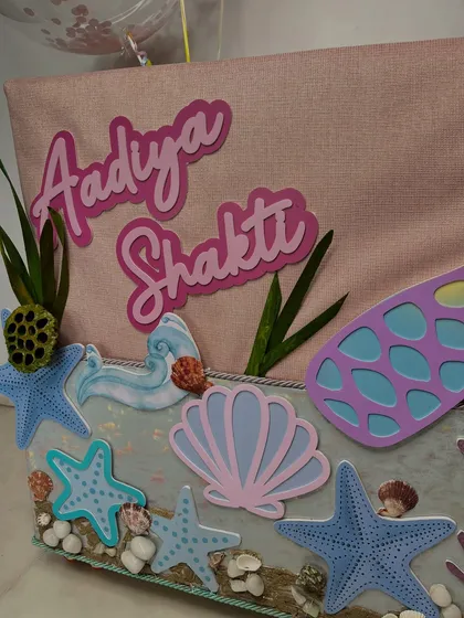 This angle shows the layering of the mermaid tail and nameplate against the pink fabric-wrapped gift box, highlighting the careful composition.