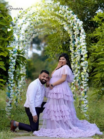 A playful and happy couple's portrait under a floral arch. The father-to-be is listening to the baby bump with a surprised expression.
