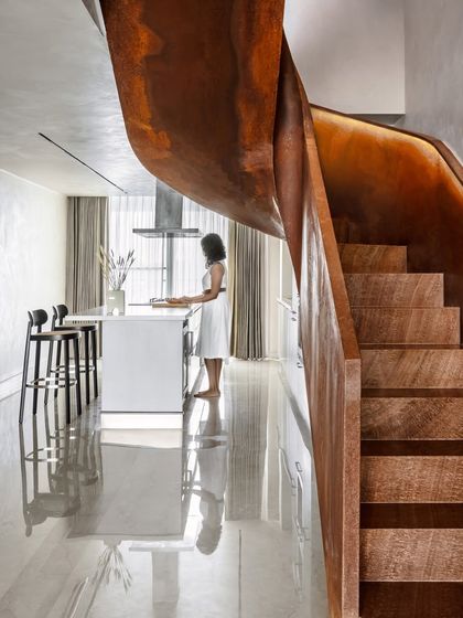 This view showcases the relationship between the sculptural staircase and the open-plan kitchen. The raw beauty of the Corten steel and wooden steps creates a powerful visual anchor in the home's otherwise understated and monochromatic design.