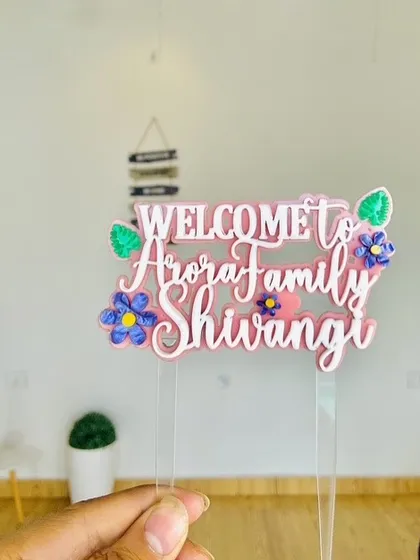 The colorful "Welcome to Arora Family Shivangi" topper held up to show its layered design and cheerful floral details.