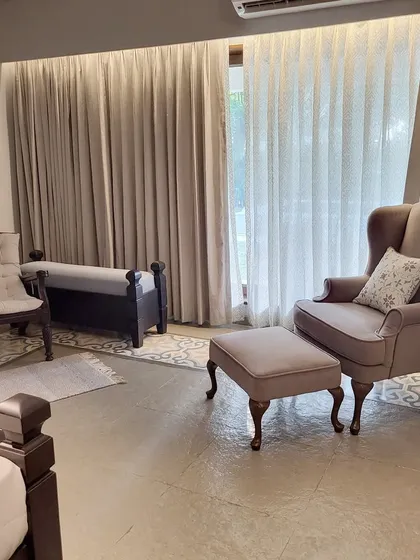 A cozy seating corner in the Alibaug villa, featuring a classic wingback chair and ottoman. The soft upholstery and flowing curtains create a serene and elegant space for relaxation.