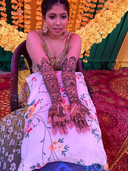 A bride seated against a festive marigold backdrop, displaying her full-arm narrative mehendi.