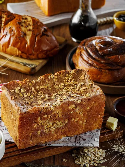 An assortment of sweet and savory breads. The focus is on the different textures and toppings, from oat-crusted loaves to cinnamon rolls, creating a rich and abundant visual for a bakery.