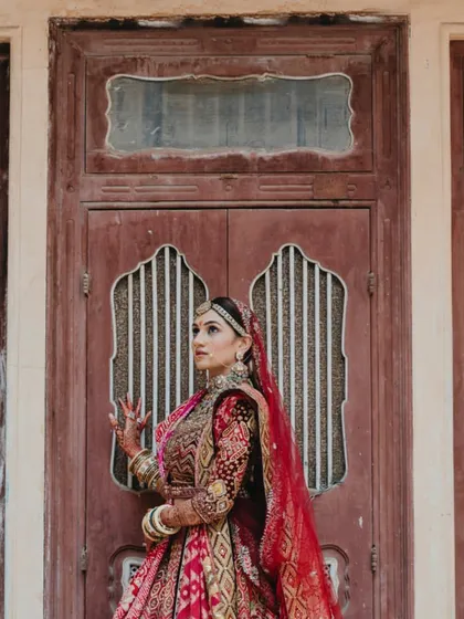 A stunning solo portrait of the bride, her intricate lehenga and jewellery shining against a rustic wooden door.