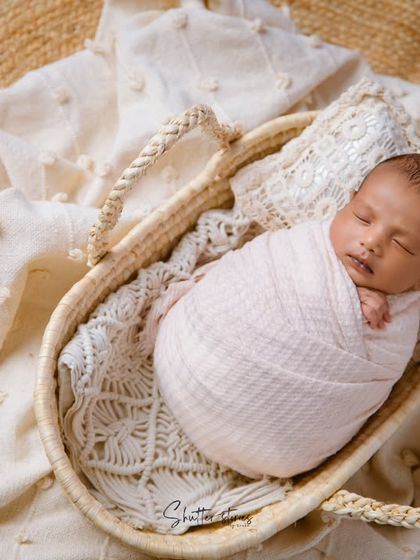 A beautifully swaddled newborn in a soft pink wrap, sleeping peacefully in a rustic basket on a textured blanket.