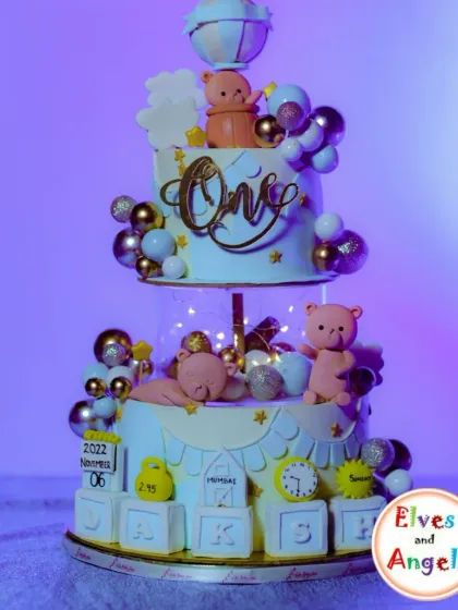 An incredibly detailed two-tier cake for a first birthday. The cake tells a story with a teddy bear, milestone details like weight and time of birth, and a hot air balloon topper.