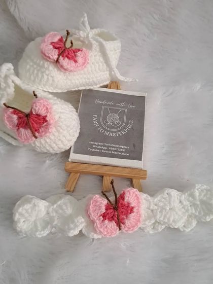 A delicate and sweet baby set featuring a butterfly theme. The white booties and matching headband are adorned with a small, pink crochet butterfly, adding a whimsical touch.