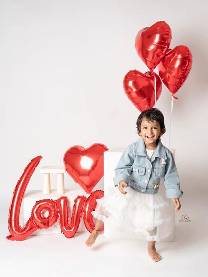 A toddler photoshoot with a 'Love' theme, perfect for Valentine's Day or any day. She poses with red heart-shaped balloons and a denim jacket, creating a sweet and modern portrait.
