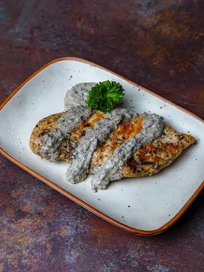 A minimalist shot of grilled chicken breast topped with a creamy mushroom sauce. The simple composition on a dark, textured plate gives it a modern and sophisticated look.