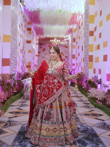 A full-length portrait of a radiant bride in her magnificent red and gold lehenga, standing in a beautifully decorated wedding hall.