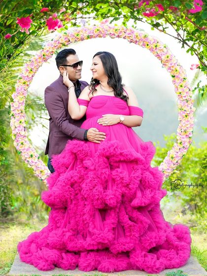 A loving couple's portrait in front of a circular floral prop. The mother-to-be's pink ruffled gown is the star of the show.