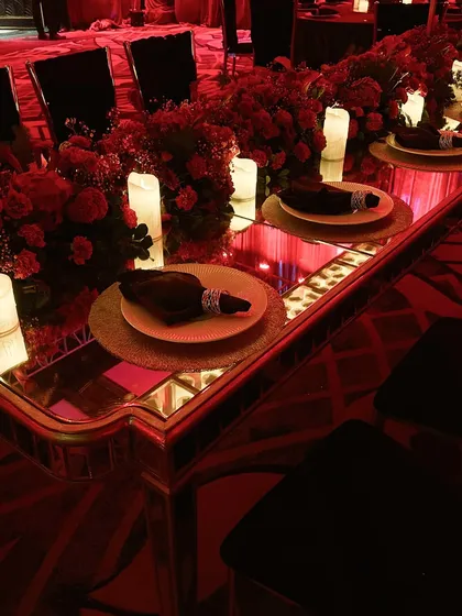 A long, mirrored dining table set for the Sangeet, with a runner of red flowers and candles, reflecting the dramatic red drapes of the ballroom.