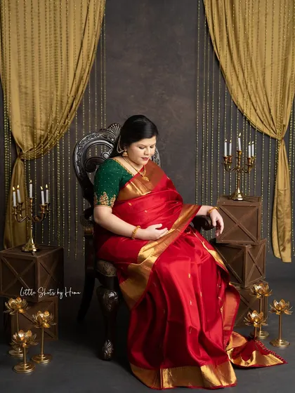 A beautiful portrait of a mother-to-be seated in a regal chair. She is wearing a rich red silk saree, looking down thoughtfully in a traditionally styled studio.