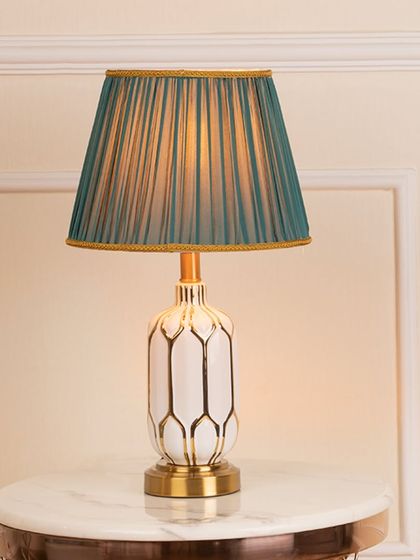 Here is the Crown Royal table lamp, shown lit on a marble side table. The warm light filters beautifully through the pleated teal shade, highlighting the elegant white and gold pattern on the ceramic base.