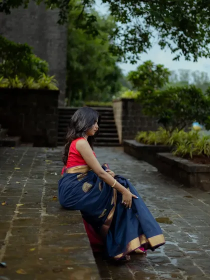 This wide shot captures the model in a quiet moment, seated on wet pavement with lush greenery in the background. It highlights the contrast between the rich saree fabric and the rustic, natural environment.