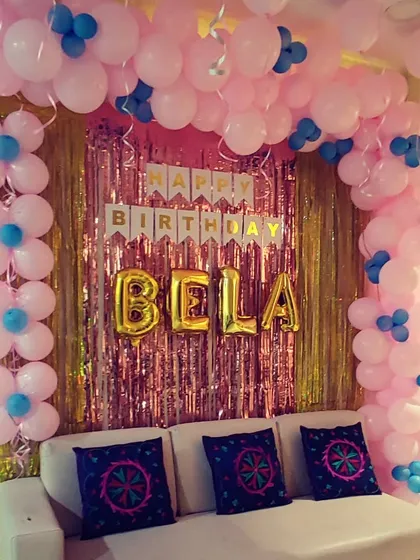 A simple birthday decoration using a pink balloon arch with blue accents. The setup frames a gold shimmer curtain and includes the birthday girl's name in large gold foil letters.