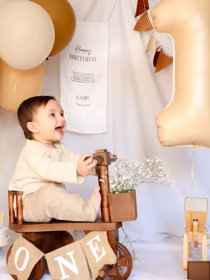 A baby boy laughs with delight while sitting on a wooden tricycle in a rustic, neutral-toned first birthday setup. This image showcases a charming and timeless theme for a milestone session.