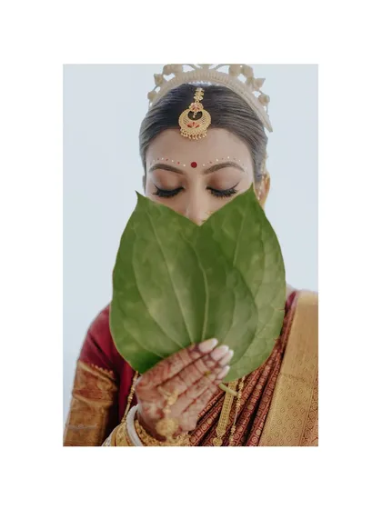A playful yet traditional pose. The bride peeks over betel leaves, her eyes telling a story of anticipation and joy, a classic moment in Bengali wedding photography.