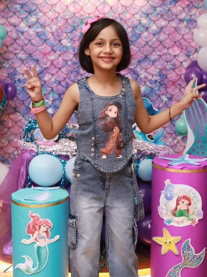 The birthday girl is all smiles, posing with a peace sign at her magical Mermaid-themed party. The detailed backdrop and decorations make for perfect photo opportunities.