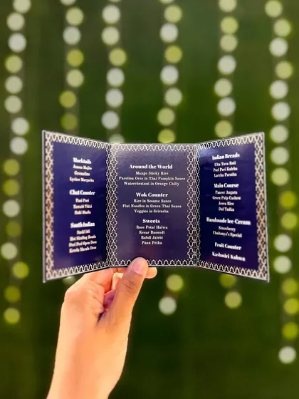A beautifully designed navy blue and gold menu card, held up against a backdrop of festive lights. The intricate design reflects the premium and detailed nature of my catering service.
