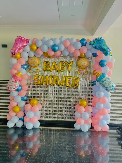 A classic baby shower backdrop with a balloon arch in pink and blue, a silver tinsel curtain, and baby-themed foil balloons.