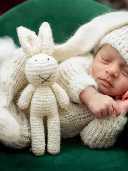 A newborn dressed in a white bunny outfit sleeps on a miniature green sofa, holding a crochet bunny.