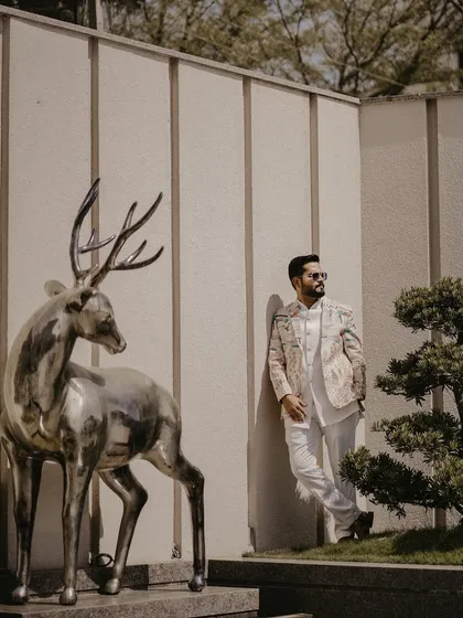 A fashionable solo portrait of the groom leaning against a wall next to a metallic deer sculpture. This shot combines modern art, architecture, and high-fashion wedding attire.