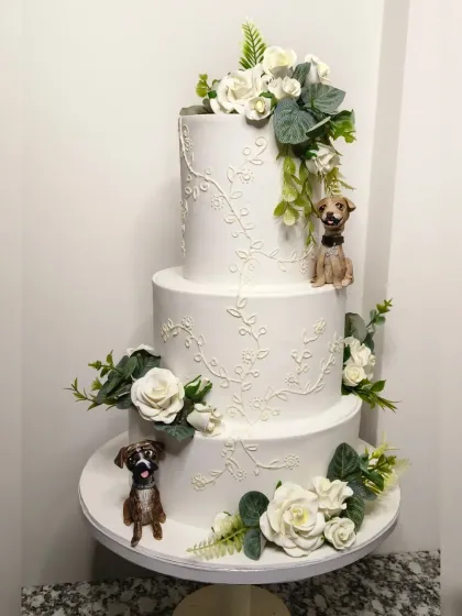 A full view of the beautiful three-tiered wedding cake featuring the couple's dogs. The clean white fondant, delicate royal icing vines, and classic sugar flowers create a perfect canvas for the personalized, hand-sculpted pet figurines.