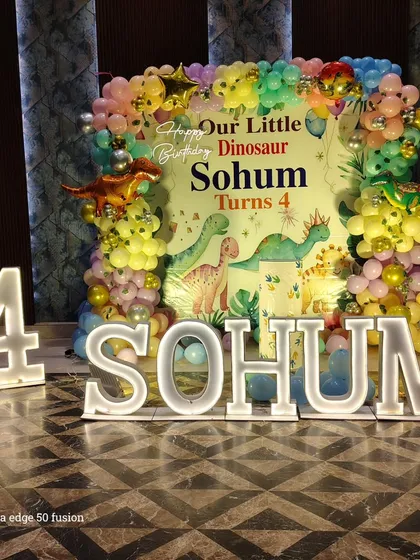 A "Little Dinosaur" themed party for a 4th birthday, featuring a custom backdrop, colorful balloon garland, and large light-up letters spelling the child's name.