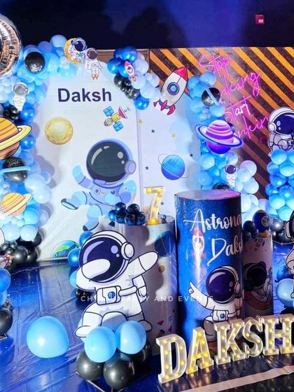 An astronaut and space-themed decoration for a 7th birthday. This detailed setup includes astronaut cutouts, planet graphics, custom pedestals, and a blue and black balloon arch to create an out-of-this-world experience.