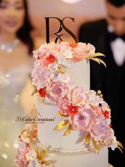 A close-up of the exquisite, handcrafted sugar flowers on a wedding cake. This shot highlights the lifelike detail of the pink peonies and red blossoms, accented with edible gold leaves and a custom monogram topper.