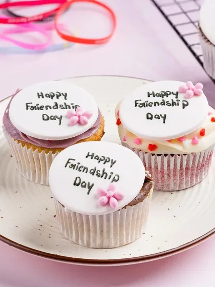 "Happy Friendship Day" cupcakes, styled with friendship bands in the background. Thematic propping like this makes the product relevant for a specific occasion.