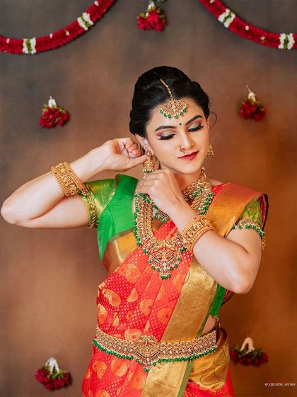 This pose, where the model adjusts her earring, feels natural and elegant. It effectively showcases the heavy jhumkas, the necklace, and the intricate details of the silk saree's border and blouse.