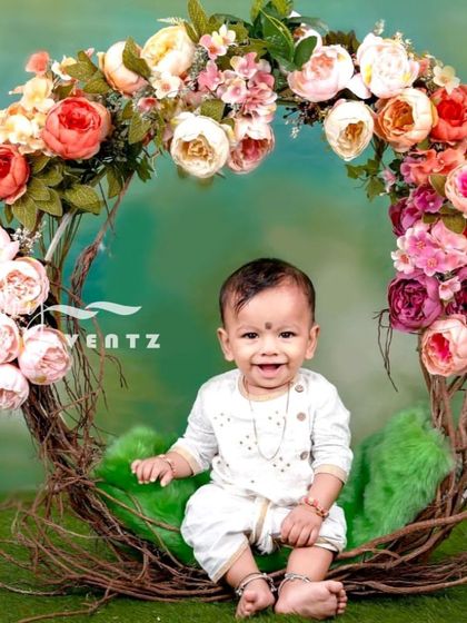 A beautiful peony-themed photoshoot for a baby boy. Dressed in a traditional white outfit, he sits happily in a rustic nest swing decorated with a large arch of pink and white peonies.