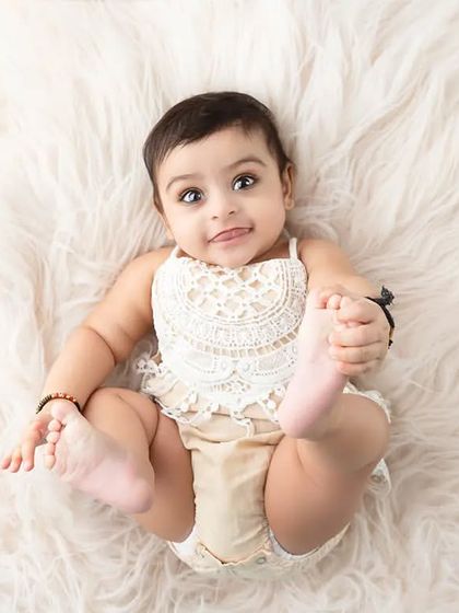 A playful baby lies on her back on a white fur rug, holding her feet.