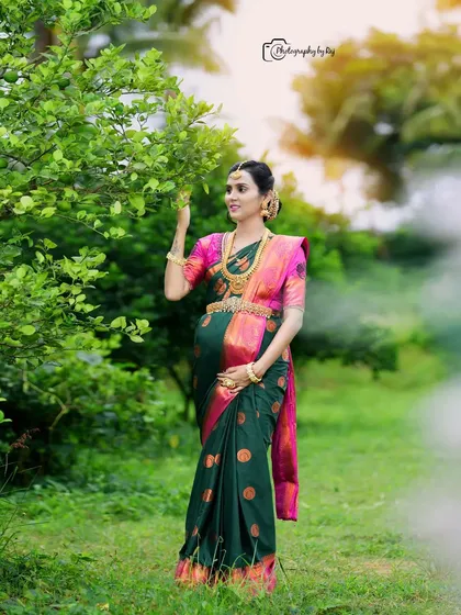 A serene outdoor portrait of a mother-to-be in a green and pink silk saree, enjoying a quiet moment in nature.