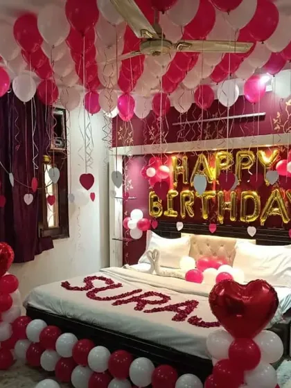 A classic romantic birthday bedroom setup. This decoration features red and white balloons on the ceiling and floor, "Happy Birthday" foil letters, and rose petals on the bed.