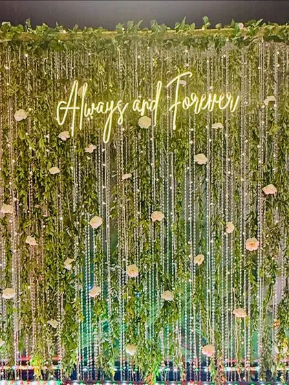 This photo backdrop brings the outdoors in with a lush wall of green foliage. I wove delicate strings of fairy lights and white roses through the leaves to create a magical, sparkling effect. The custom "Always and Forever" neon sign adds a personal and romantic touch, making it an ideal spot for capturing memories.
