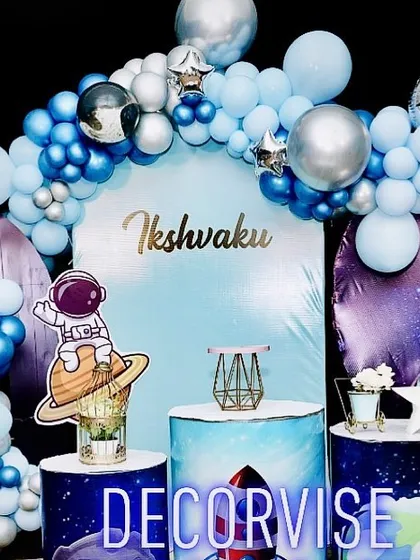 We can launch your child's party into another galaxy with a custom space theme. This cosmic setup for Ikshvaku includes a galaxy-print backdrop, an astronaut cutout, planet-themed pedestals, and a metallic blue and silver balloon arch to create an out-of-this-world experience.