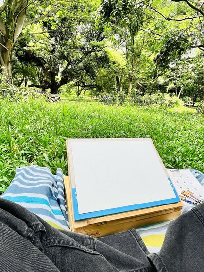 My setup for the day. A comfy blanket, a blank page on my easel, and the endless inspiration of Cubbon Park's greenery in front of me. This is how every great painting begins.