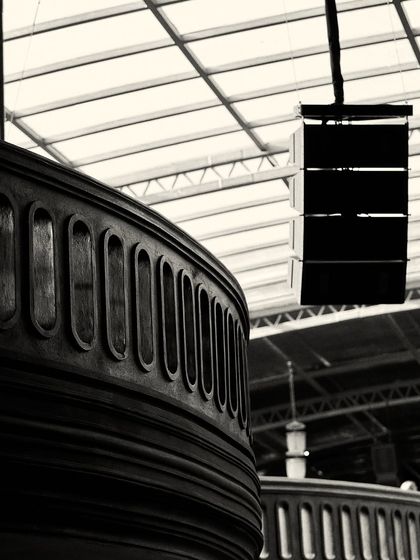 A close-up of the carved wooden balustrade, showing its texture and repeating pattern. The industrial speaker and glass ceiling provide a modern contrast.