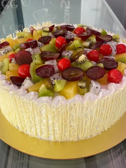 A beautiful mixed fruit gateaux, generously topped with fresh kiwi, grapes, dragon fruit, and cherries, all surrounded by a white chocolate cage.