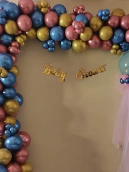 A baby shower balloon arch in a striking color combination of metallic pink, blue, and gold. The 'Baby Shower' banner hangs in the center, creating a festive frame for the celebration.