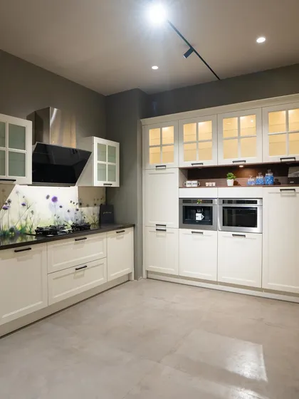 A classic white kitchen from our Coimbatore showroom. Glass-fronted upper cabinets with internal lighting and a floral backsplash add a touch of traditional charm and personality.