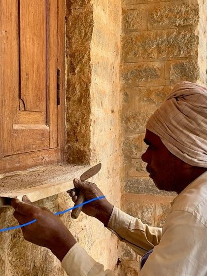 A craftsman applies mortar to the base of a restored wooden window frame, sealing it against the stone wall. This careful finishing work is essential for weatherproofing and longevity.