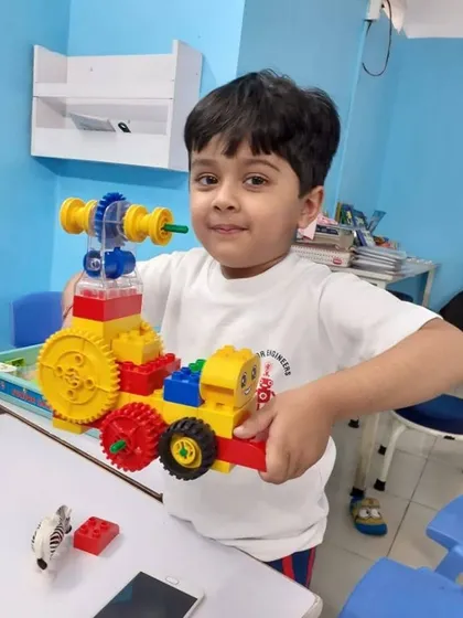 A student proudly holds up his LEGO vehicle, which features large wheels and a complex gear system he assembled.