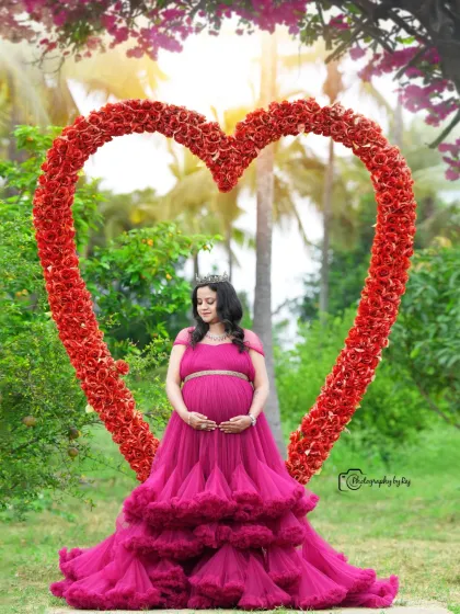 A regal portrait of a mother-to-be in a wine-colored ruffled gown and crown, framed by our red floral heart prop.