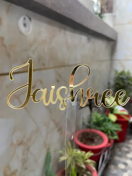 A "Jaishree" name topper in a flowing cursive font, held up to showcase its delicate design and golden shine.